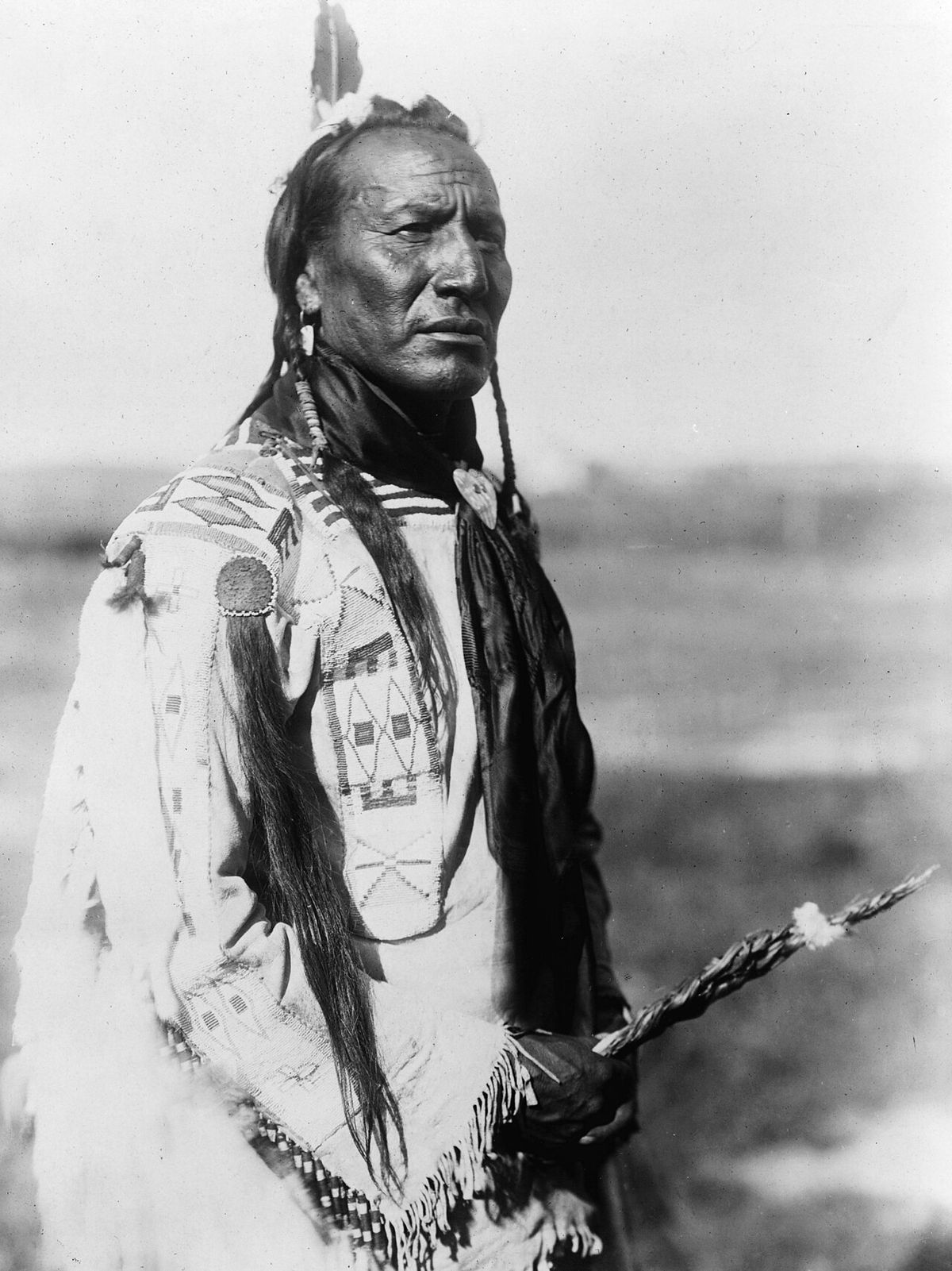 Native American Peigan Big Mouth Spring, three-quarter length portrait, standing, facing right, braids, one feather, beaded buckskin shirt, 